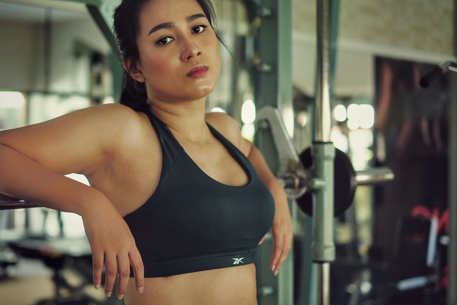 The image depicts a young woman with medium skin tone and dark hair pulled back, standing in a gym environment. She is wearing a black athletic sports bra with a subtle logo on the front, revealing he